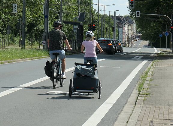 Zwei Fahrradfahrende auf einem Radweg. An einem Fahrrad ist ein Anhänger befestigt. Im Hintergrund Autos auf einem eigenen Fahrstreifen.