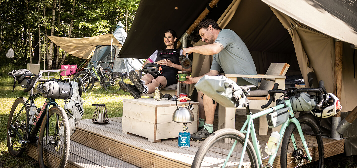 Zwei Menschen mit Gravelbikes sitzen im Eingangsbereich auf einer Holzveranda vor einem großen Zelt.