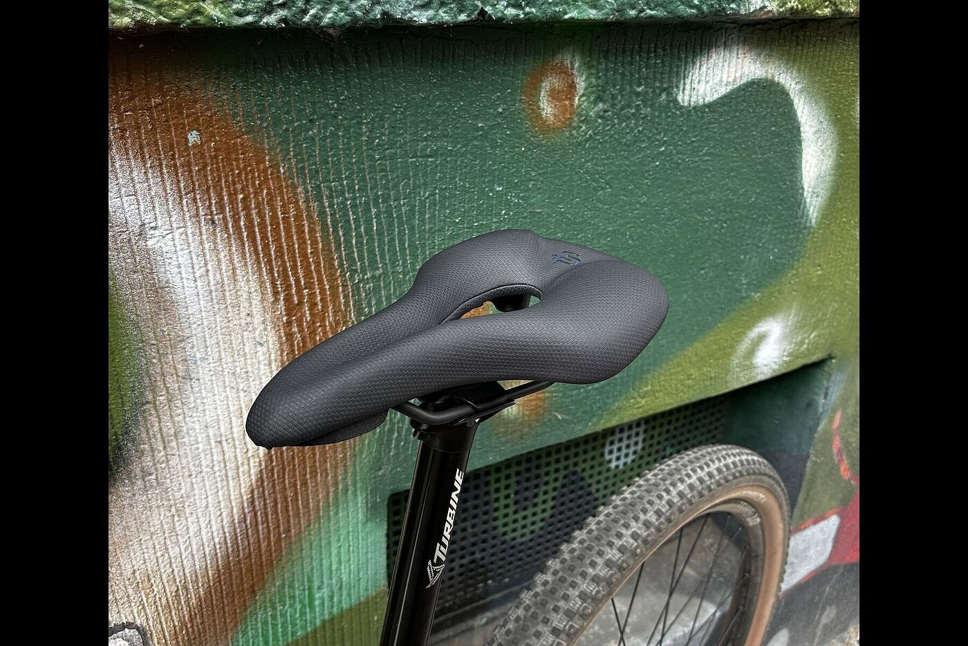 Schwarzer Fahrradsattel mit mittlerer Aussparung, montiert an einem Fahrrad vor einer bemalten Wand.