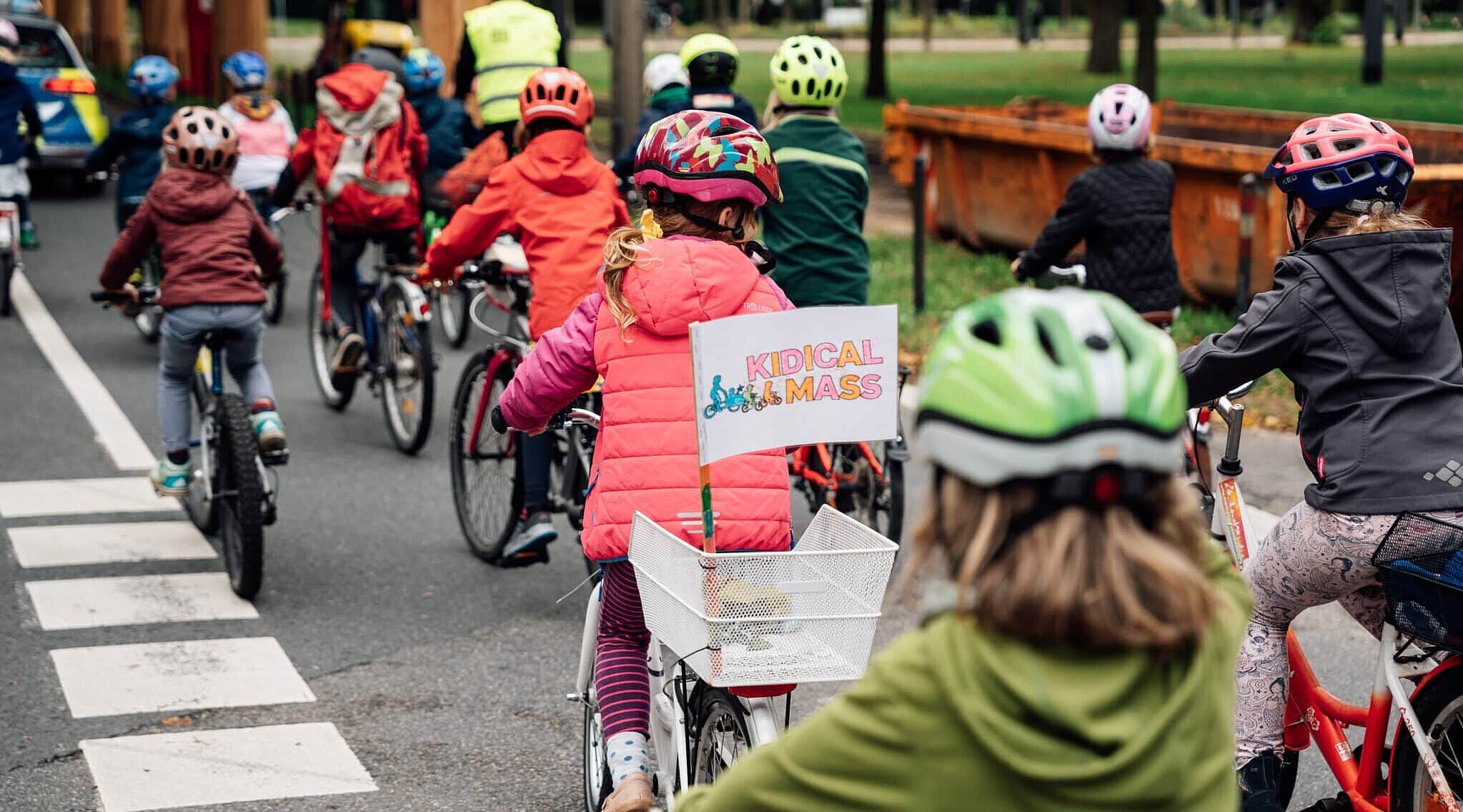 Mehrere Kinder und Erwachsene fahren bei einer Kidical Mass gemeinsam mit Fahrrädern und Helmen auf einer Straße. Ein Kind trägt eine Weste und hat ein Schild mit „Kidical Mass“ am Fahrradkorb.